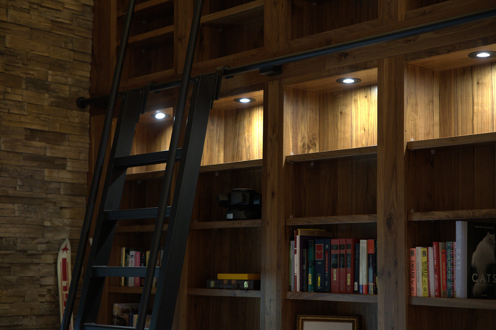 Built-in wood library bookshelf with recessed shelf lights and a matte black rolling ladder mounted on a rail, wired by iPowerDenver
